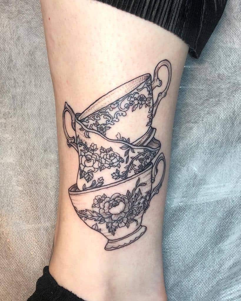 Black and grey tattoo of three stacked vintage teacups, each featuring intricate floral patterns, creating a delicate and elegant design