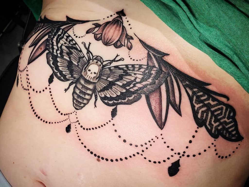 Black and grey chandelier tattoo featuring a death’s-head hawk moth, delicate bead-like chains, floral accents, and bold shading