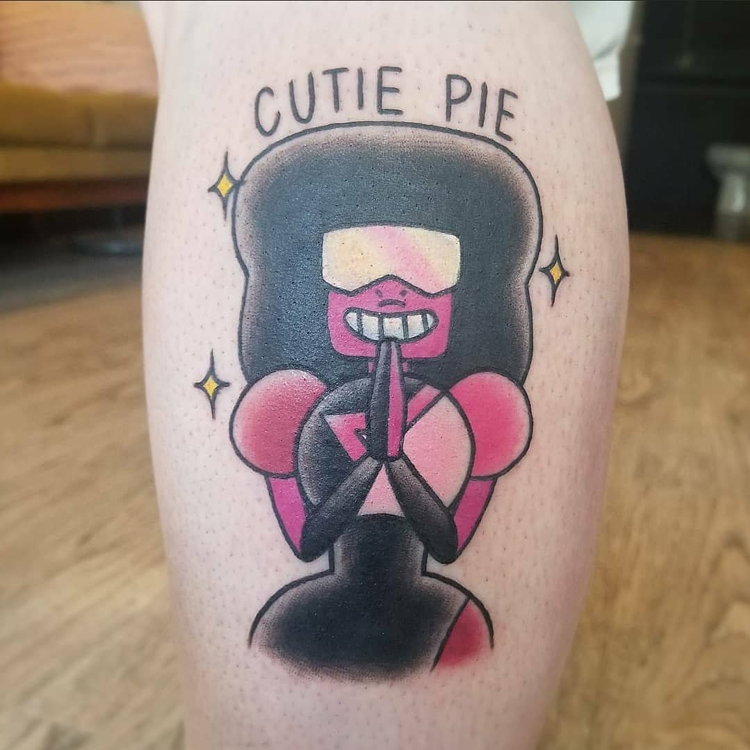 Playful tattoo of Garnet from Steven Universe with a wide grin and the words 'Cutie Pie' above her head, surrounded by small sparkles, on the leg