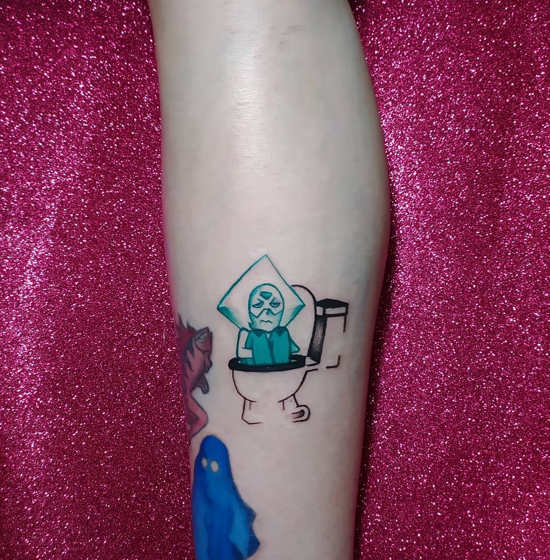 Humorous tattoo of Peridot from Steven Universe sitting on a toilet, with an annoyed expression, on the forearm