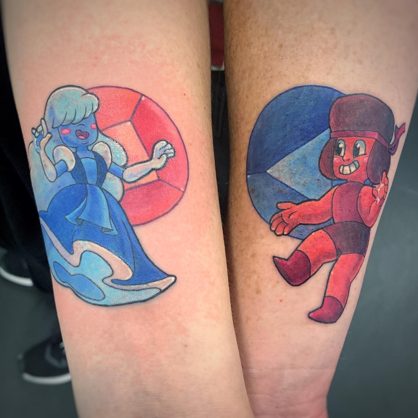 Matching tattoos of Ruby and Sapphire from Steven Universe on forearms, with Ruby in red and Sapphire in blue