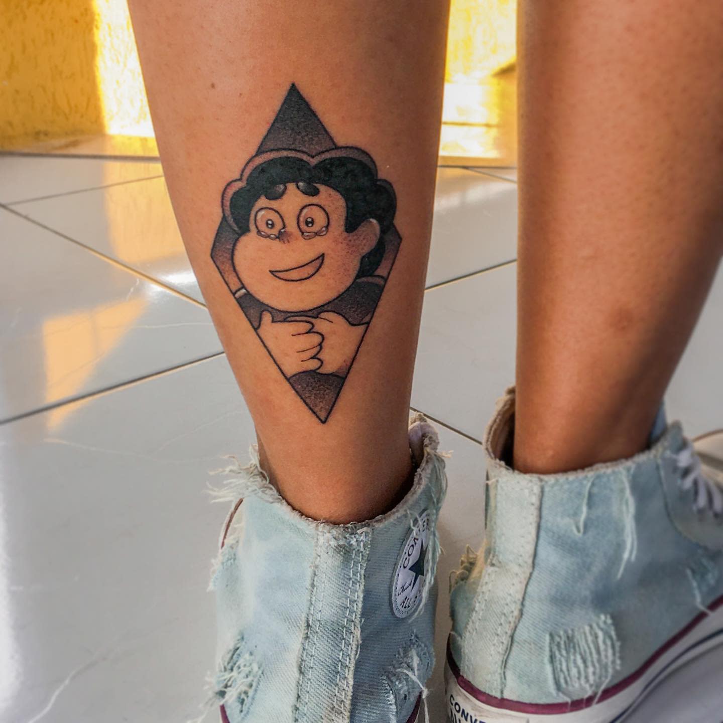 A minimalistic Steven Universe tattoo with a geometric diamond shape, showcasing a smiling Steven giving a thumbs up, placed on the lower leg