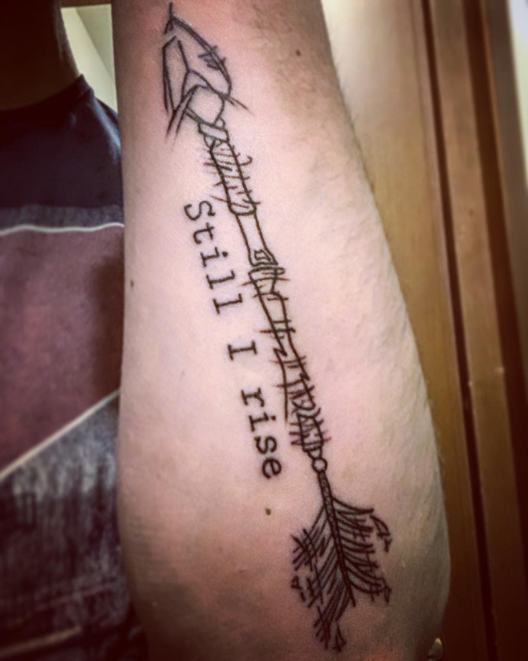 A tattoo on an arm showing an arrow with the phrase "Still I rise" along its length