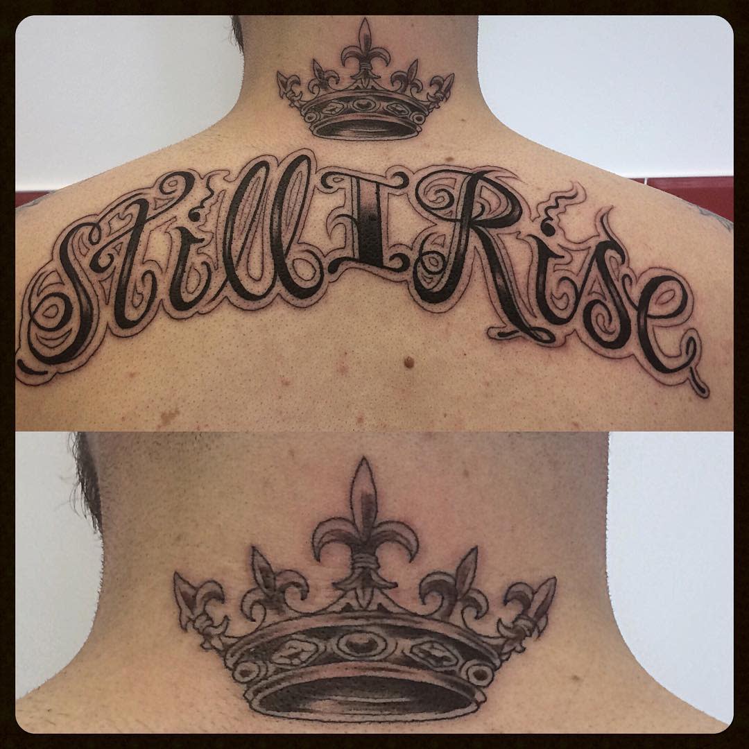 A tattoo located on the upper back features the phrase "Still I Rise" with an image of a crown positioned above the text on the nape of the neck