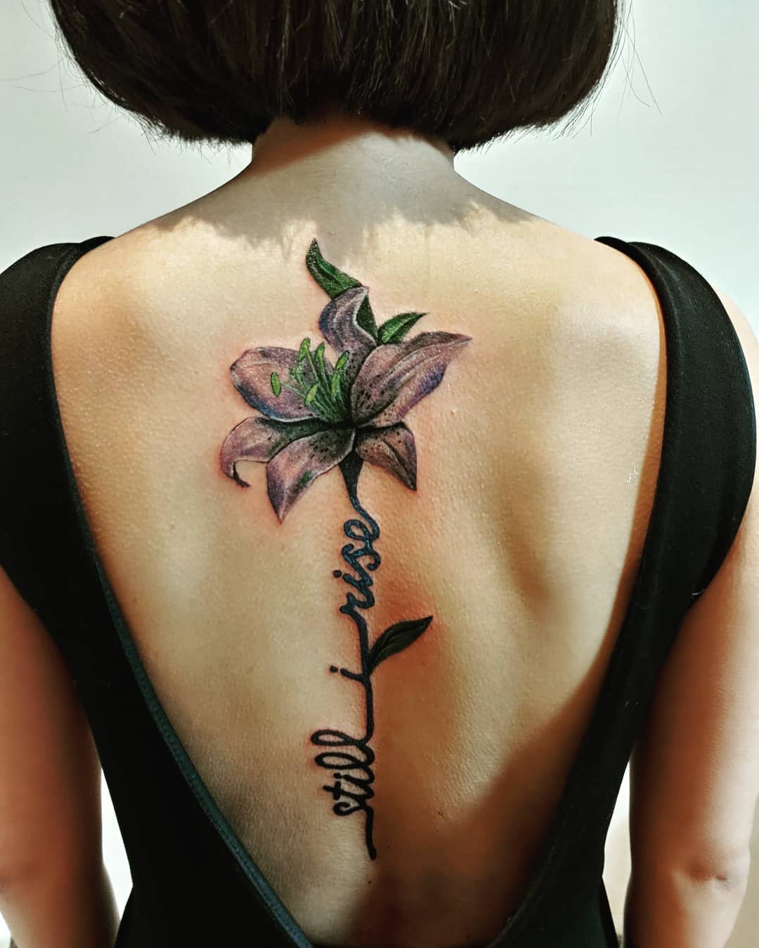 Back tattoo of blooming lily with "still I rise" text, visible through black dress with low back