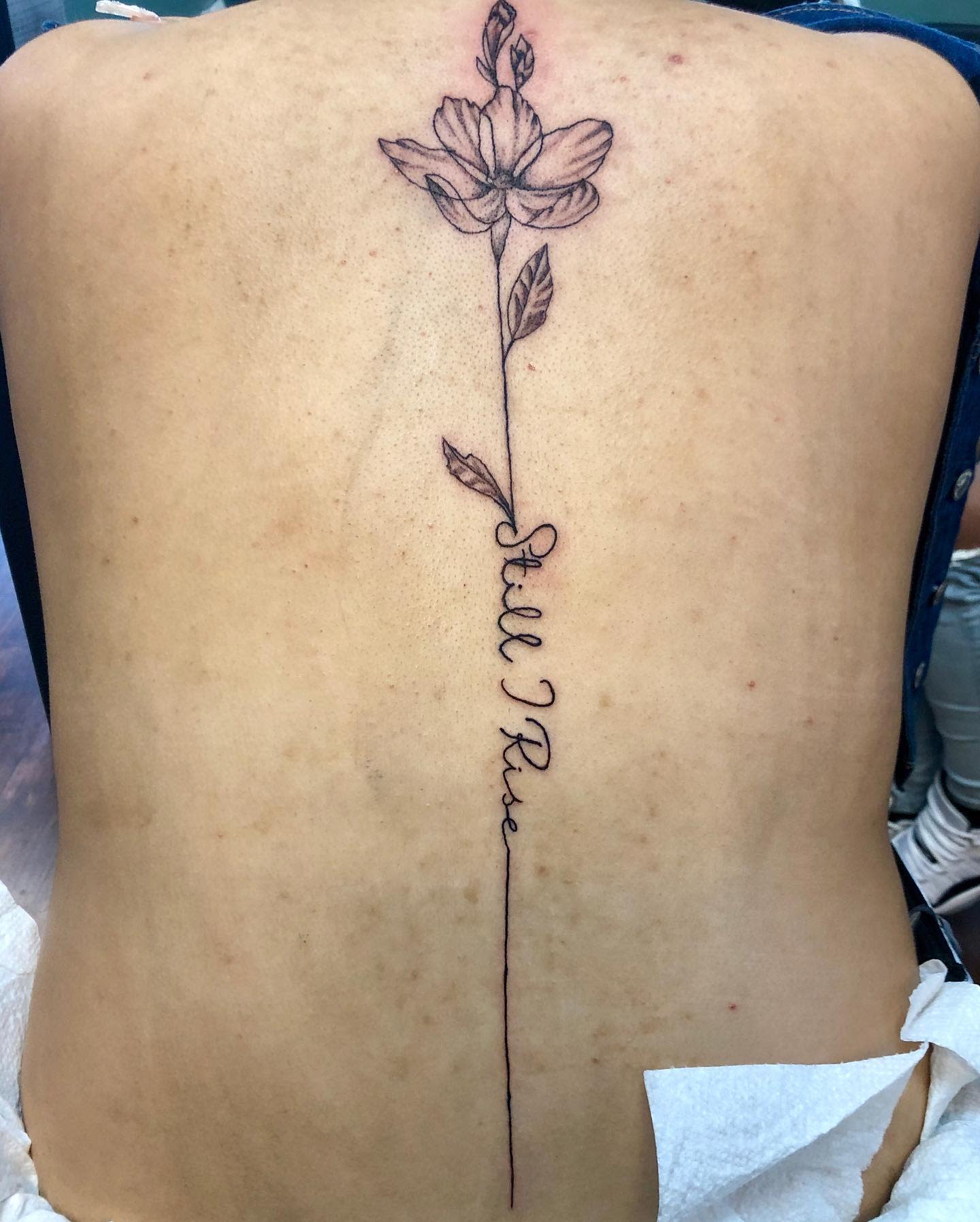 Back tattoo featuring a long-stemmed flower with "Still I Rise" inscribed along the stem