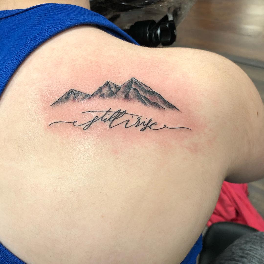Mountain tattoo with text "still i rise" on upper back, near the right shoulder