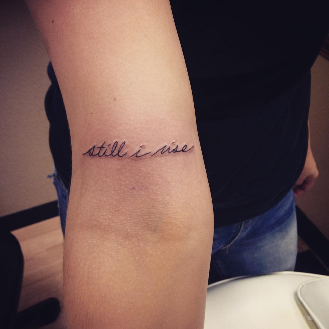 Tattoo with the phrase "still I rise" in cursive on the inside of an arm above the elbow