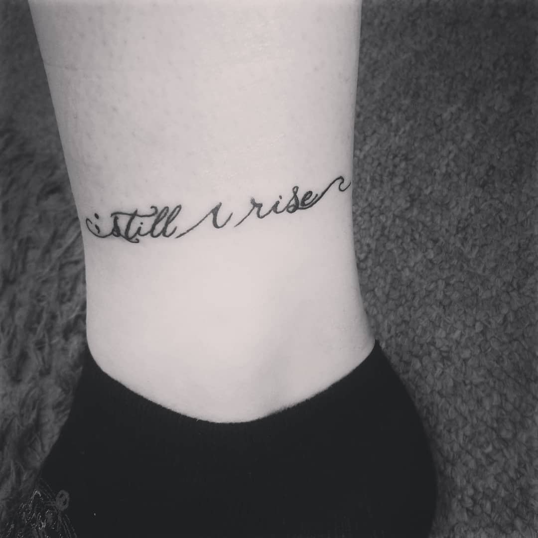Minimalist ankle tattoo reading "still I rise" in elegant cursive script