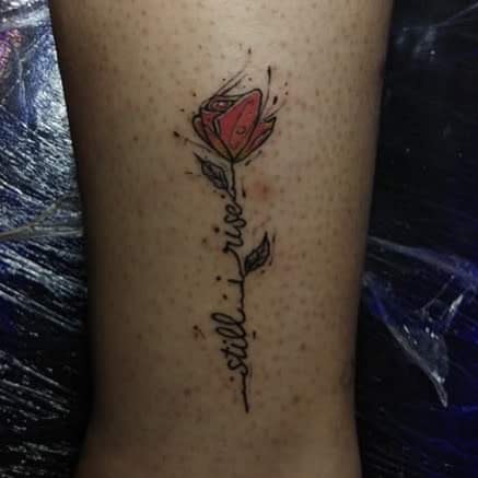 A tattoo of a red rose with the words "still I rise" along the stem on their skin