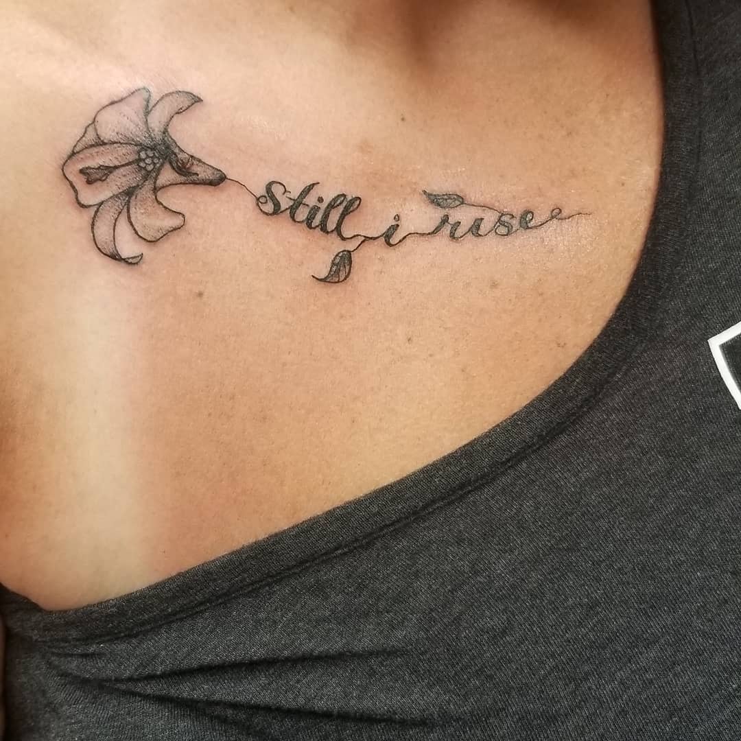 Shoulder tattoo of a flower with text "Still I Rise" on upper chest near the collarbone