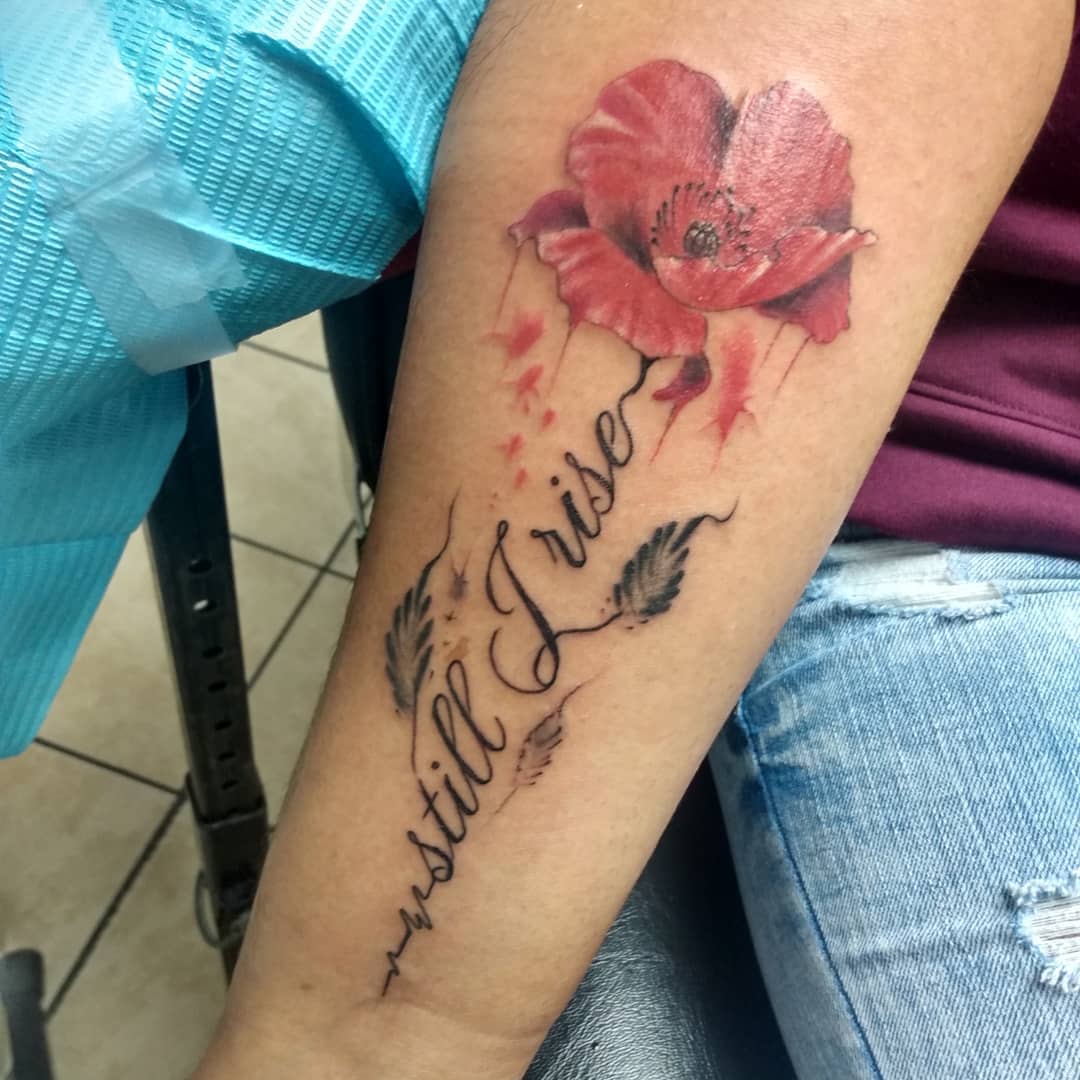 Tattoo of a red poppy with "still I rise" text and leaves on a forearm