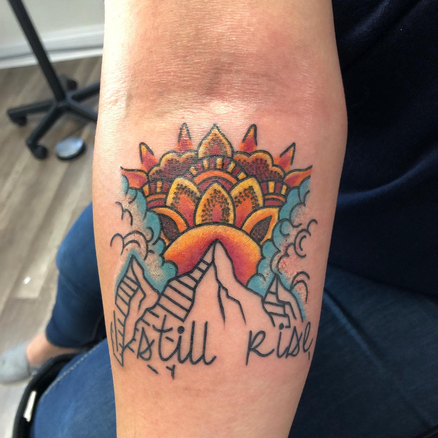 Colorful tattoo of mountains, sunrise, mandala, clouds, and text reading "I still rise" inked on an arm