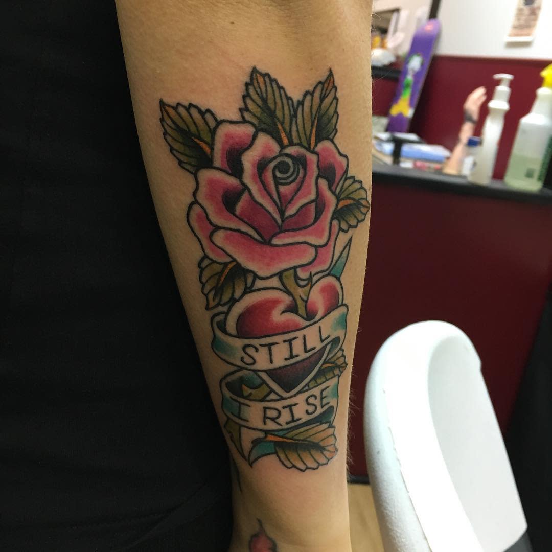 A tattoo of a rose with a banner that reads "STILL I RISE" on an arm