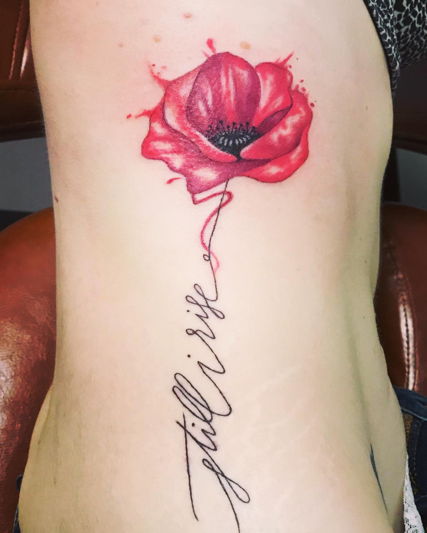 Tattoo of a red poppy flower and cursive text "still I rise" on their side torso