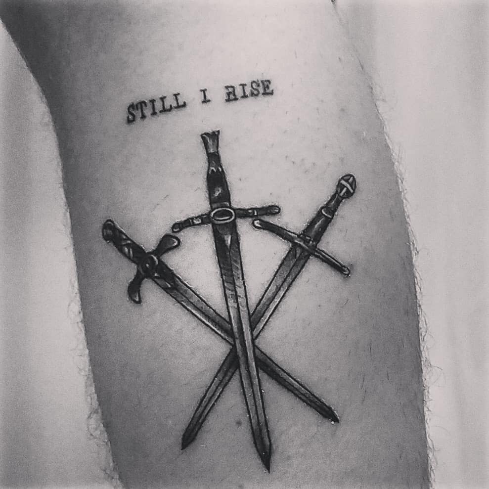 Tattoo of three crossed swords below the text "STILL I RISE" on a leg