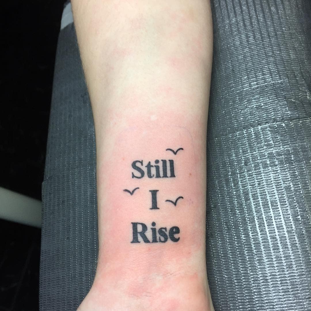 Wrist tattoo with the text "Still I Rise" flanked by small birds on each side
