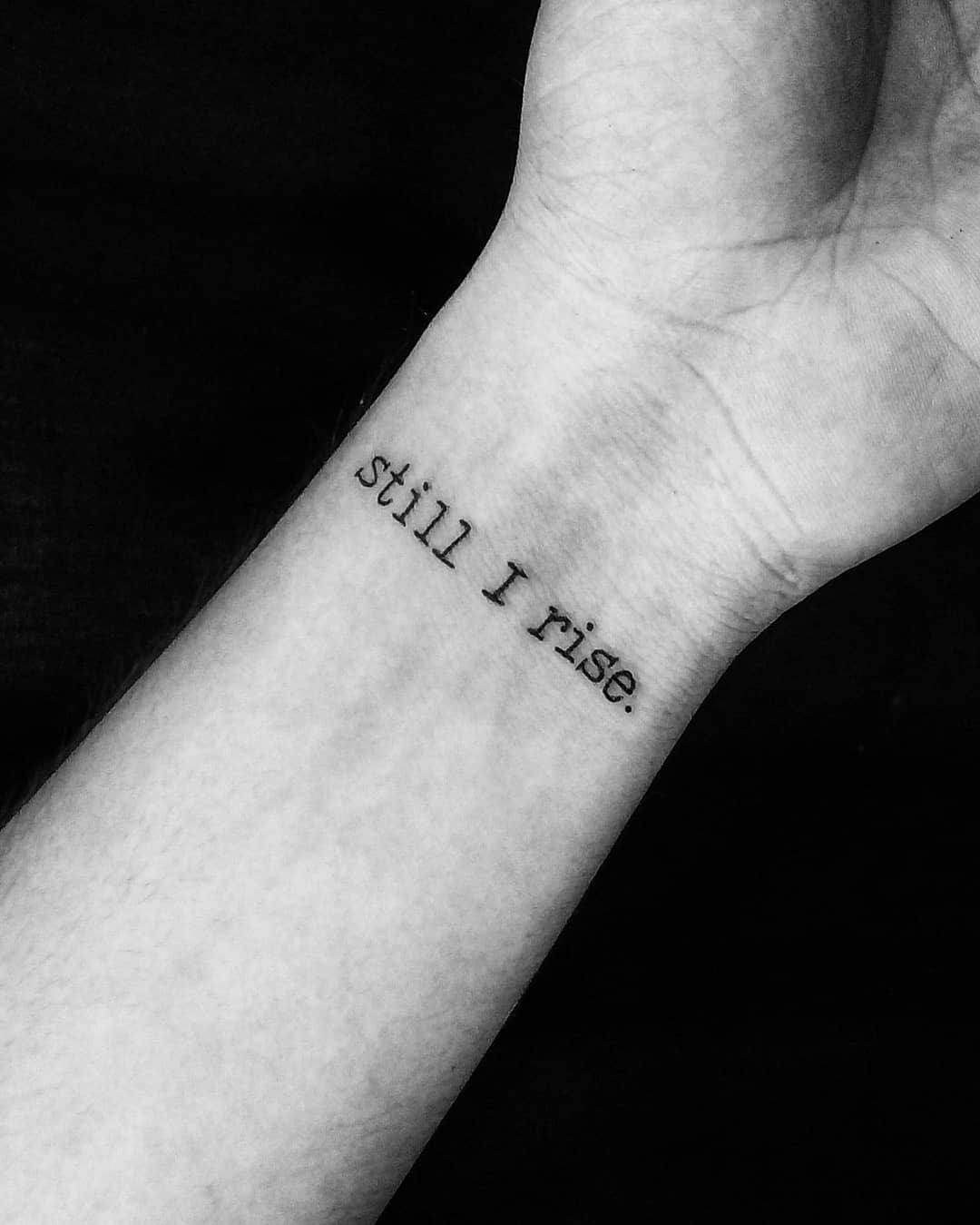 A wrist tattoo with the words "still I rise" written in a typewriter font