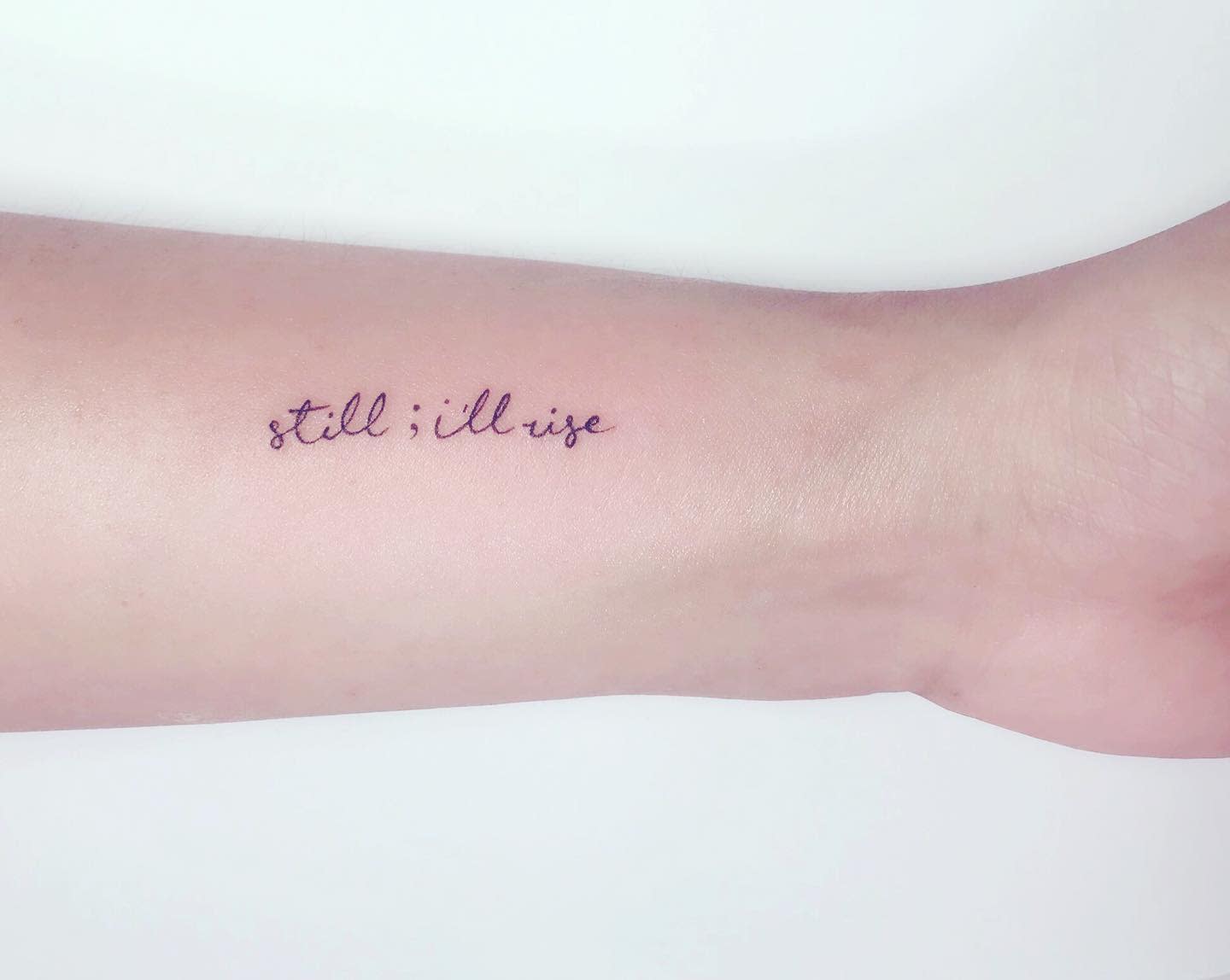 A forearm tattoo reads "still; i'll rise" in black cursive script