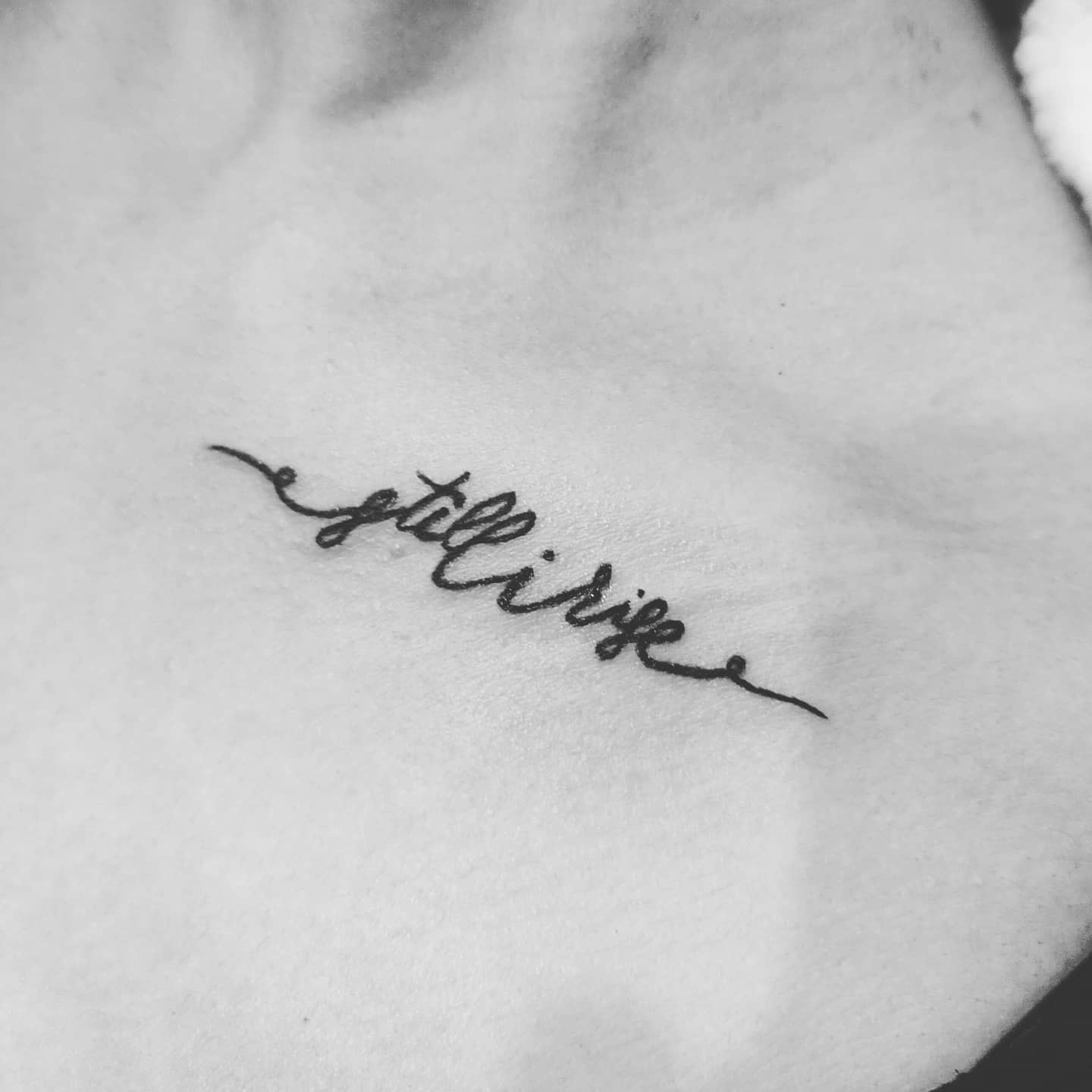 Black and white tattoo that reads "still i rise" in cursive script on skin