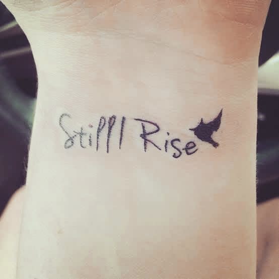 Tattoo on wrist reads "Still I Rise" with a small bird silhouette at the end