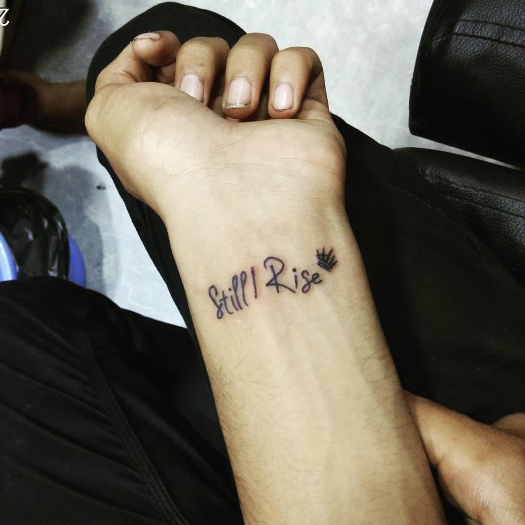 A wrist tattoo reads "Still I Rise" with a small crown symbol at the end