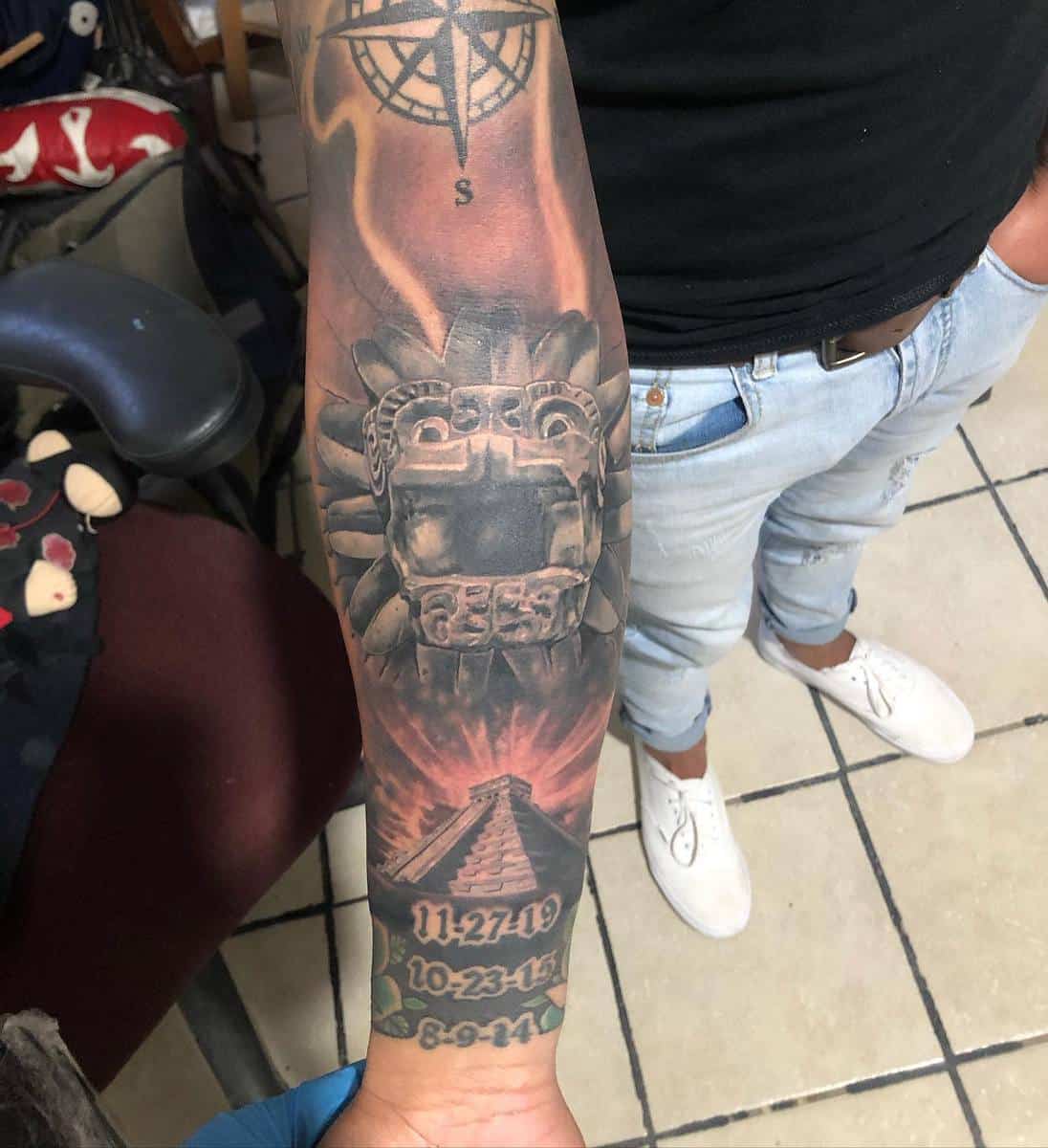 Forearm tattoo of Quetzalcoatl with a stone-carved head, Chichen Itza pyramid, and date inscriptions