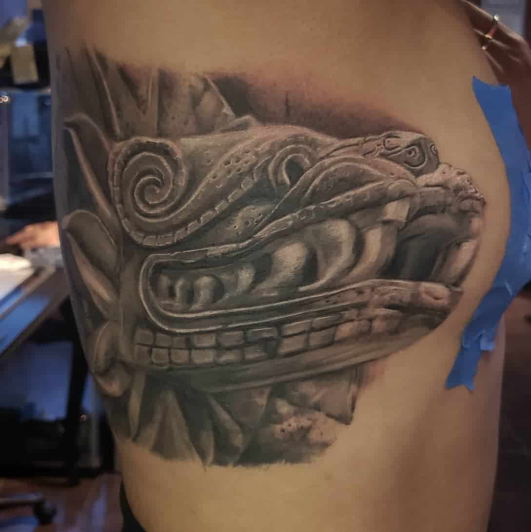Detailed side tattoo of Quetzalcoatl's stone-carved head, featuring bold shading and realism