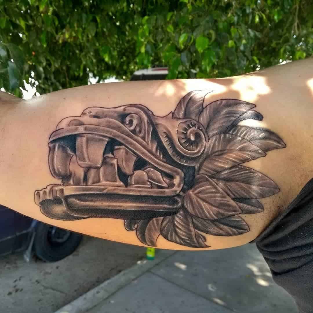 Realistic bicep tattoo of Quetzalcoatl's stone-carved head with detailed featherwork