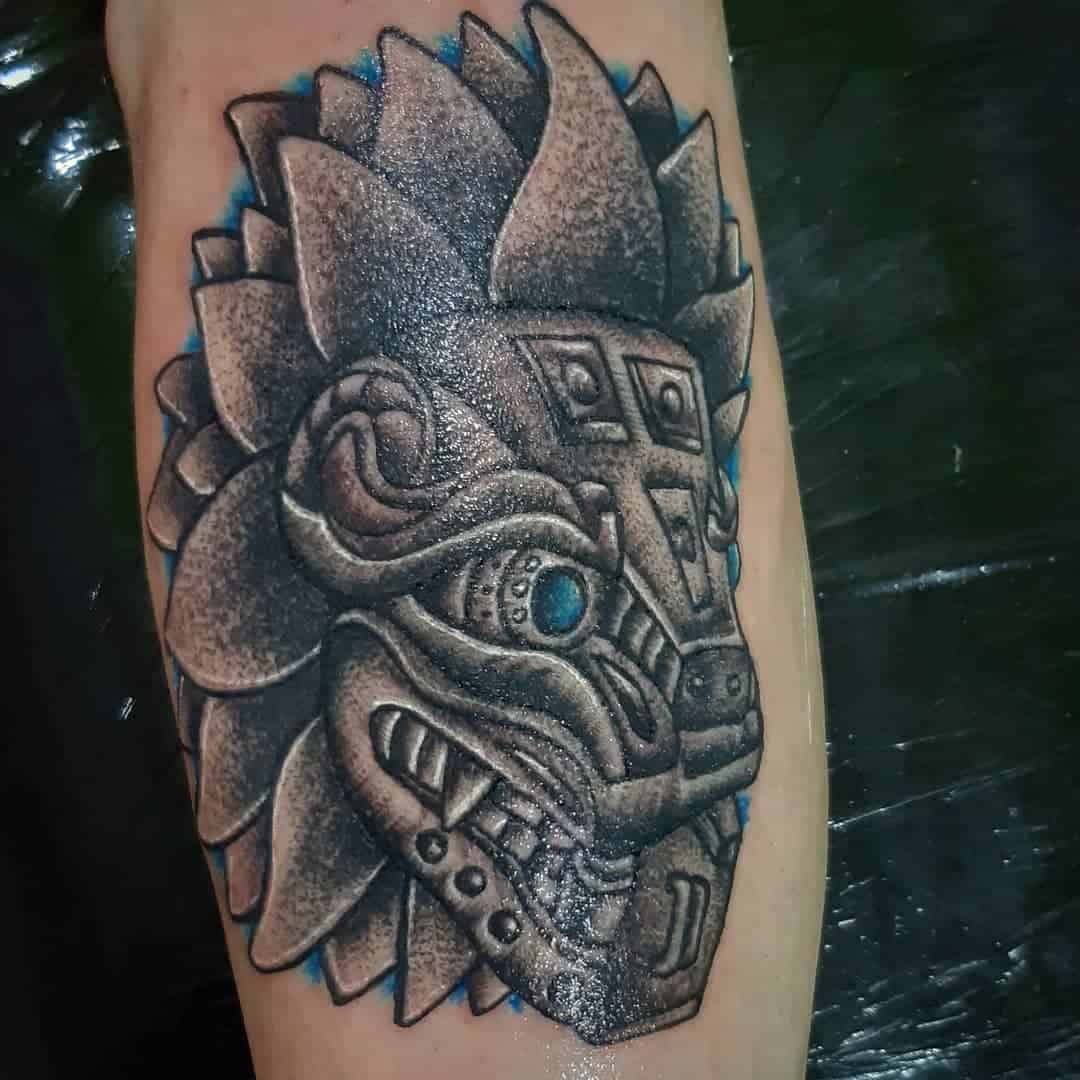 Stone-carved Quetzalcoatl head tattoo with textured shading and glowing blue eye accents