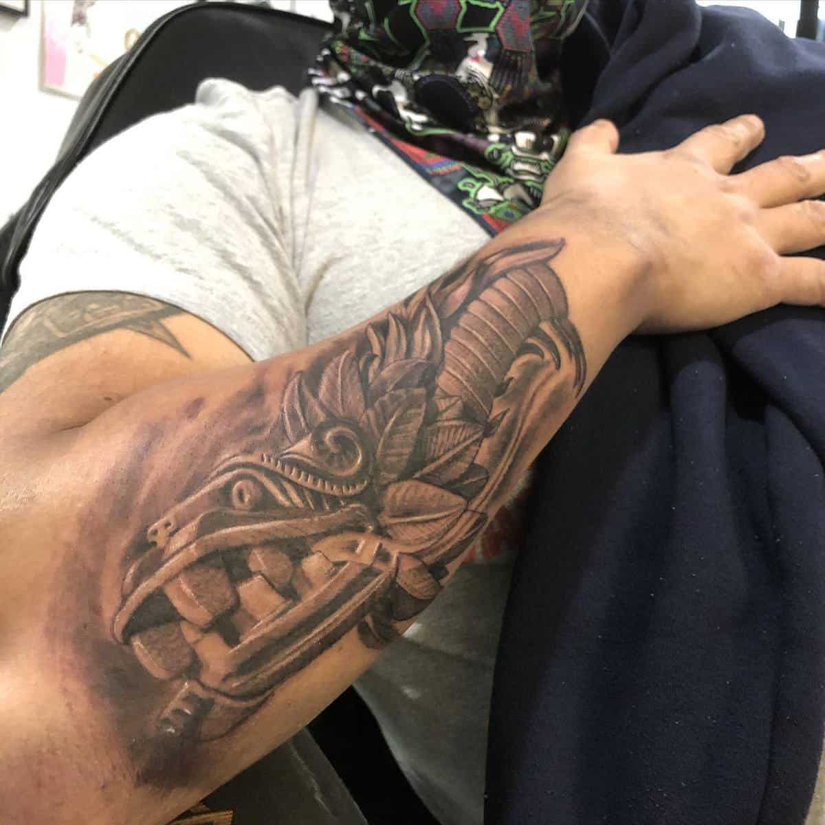 Forearm tattoo of Quetzalcoatl's stone-carved head with intricate feather details and shading