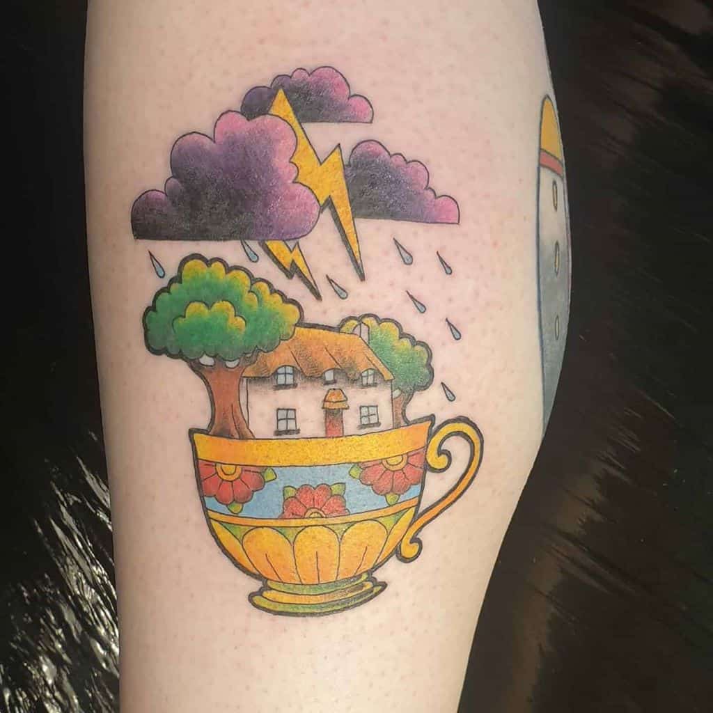 Colorful tattoo of a cottage and trees inside a floral teacup with a storm cloud, lightning, and rain above it