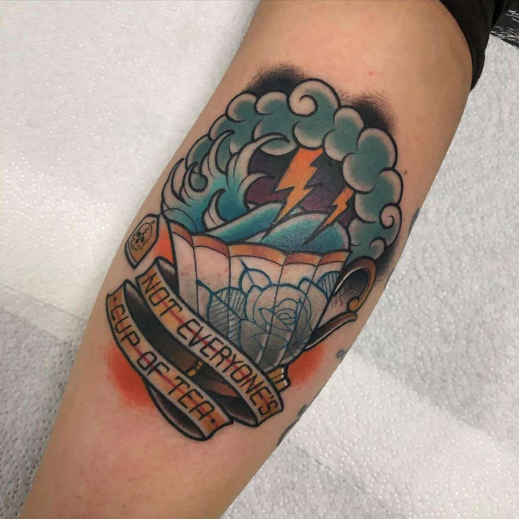 Colorful tattoo of a blue floral teacup with a storm inside, featuring lightning and waves, with a banner reading "Not Everyone's Cup of Tea" below and a tea bag with a skull