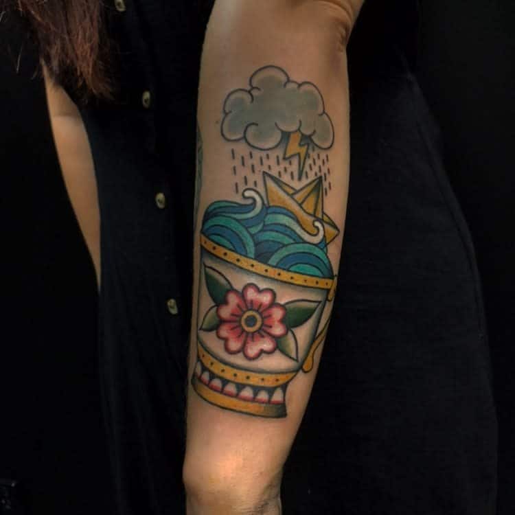 Colorful tattoo of a floral teacup with ocean waves and a paper boat sailing under a storm cloud with lightning