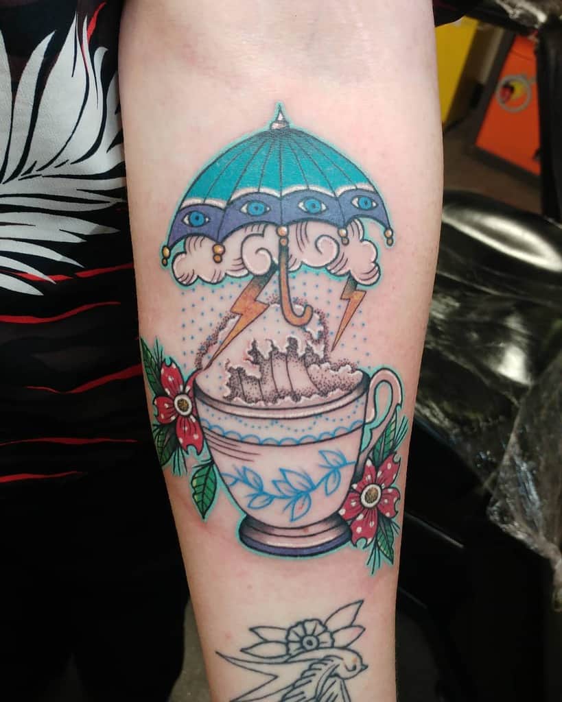 Colorful tattoo of a blue floral teacup with big waves, clouds and lightning under a blue umbrella with eyes, surrounded by red flowers and leaves