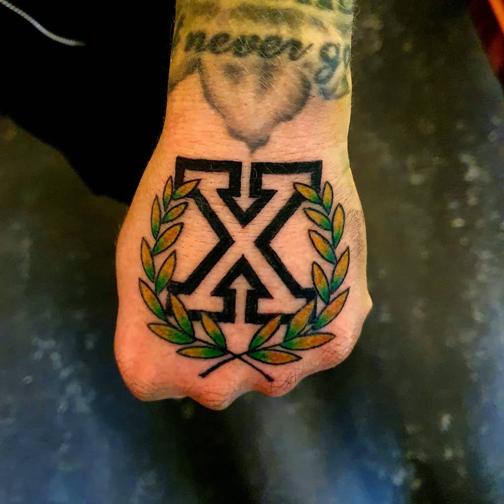 Bold hand tattoo featuring a black straight edge 'X' with arrow details, framed by a green laurel wreath