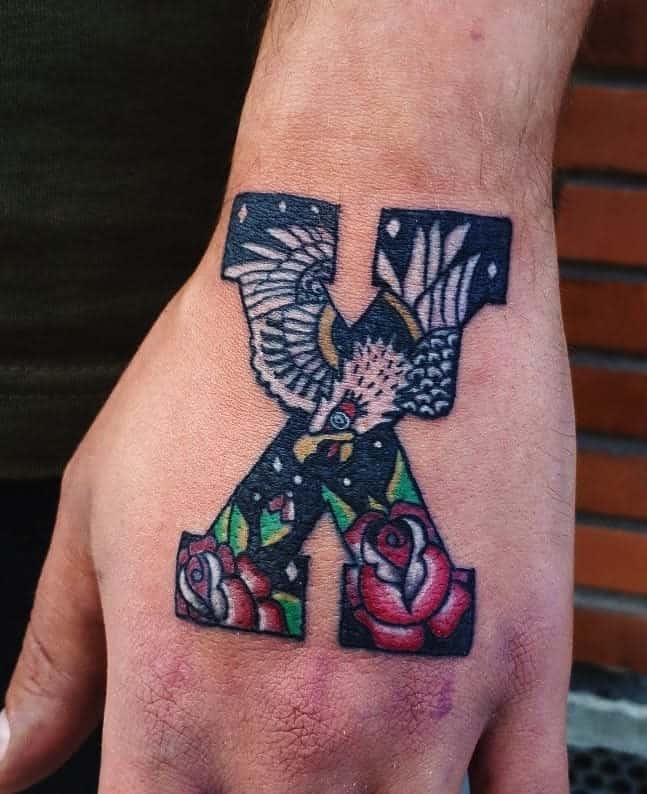 Hand tattoo featuring a bold straight edge 'X' filled with a detailed eagle, red roses, and green leaves