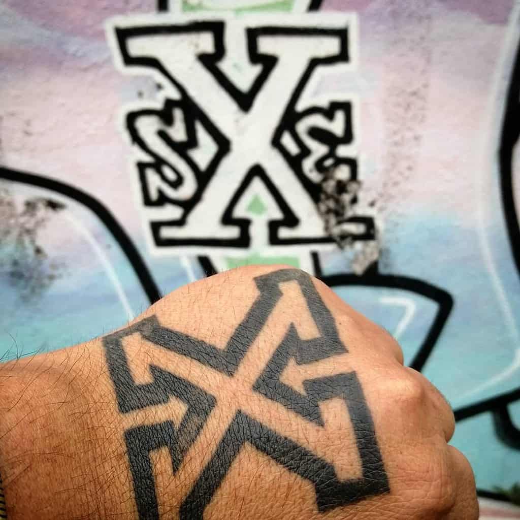 Bold black straight edge 'X' hand tattoo with sharp geometric lines, matching a graffiti-style 'SXE' design in the background