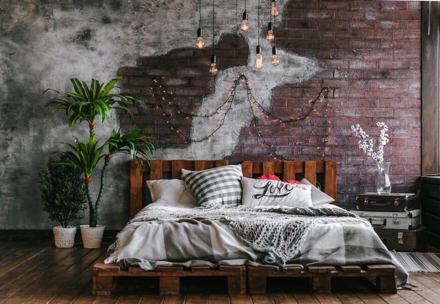 Cozy bedroom with a pallet bed, plaid pillows, and string lights on a brick wall; plants and suitcases are nearby, adding a rustic touch