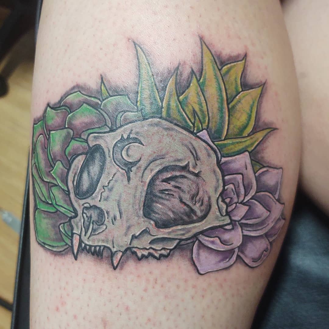 Tattoo of a cat skull surrounded by green and purple succulent plants on skin