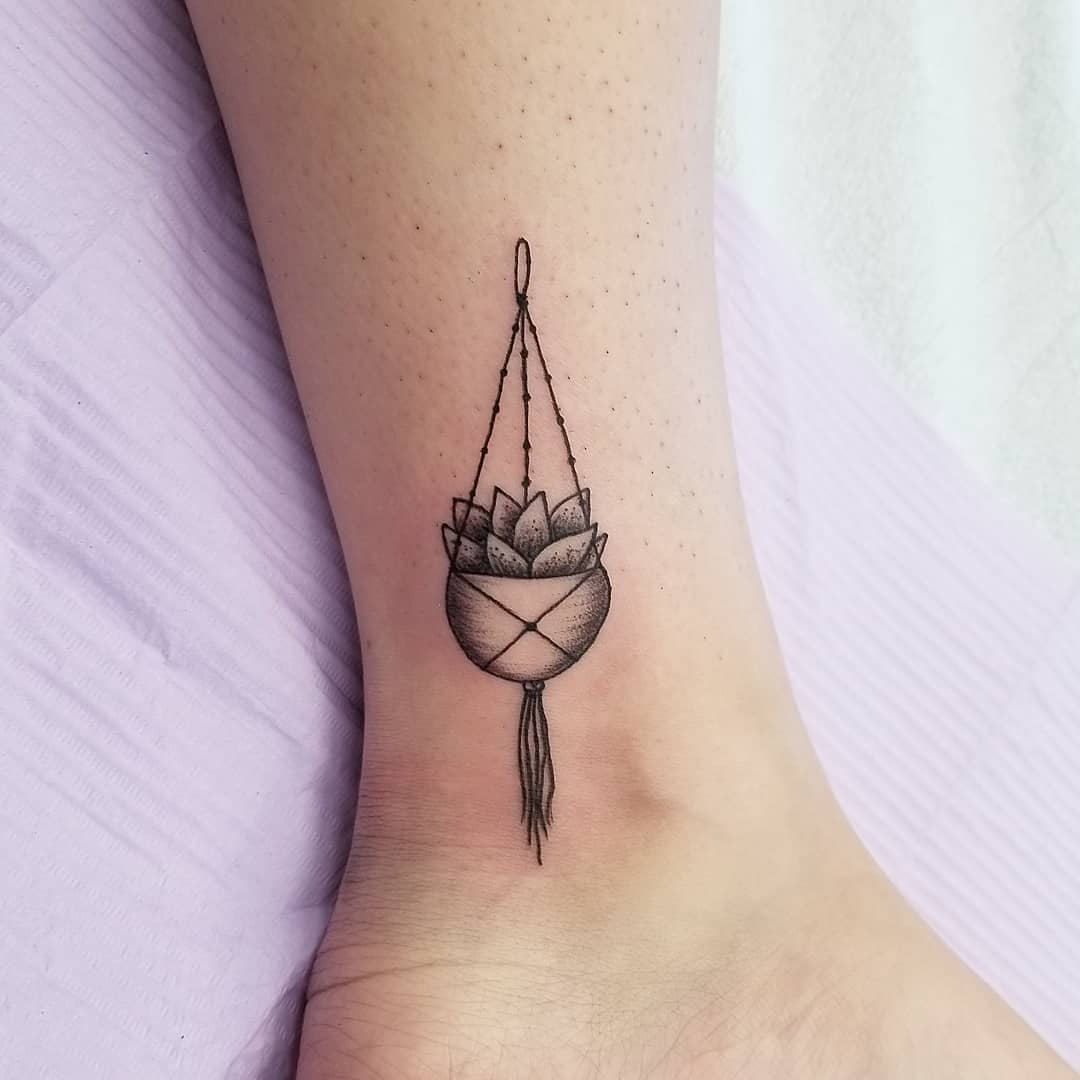 A tattoo of a hanging potted plant on the inner ankle, with the pot featuring a macramé design and a tassel