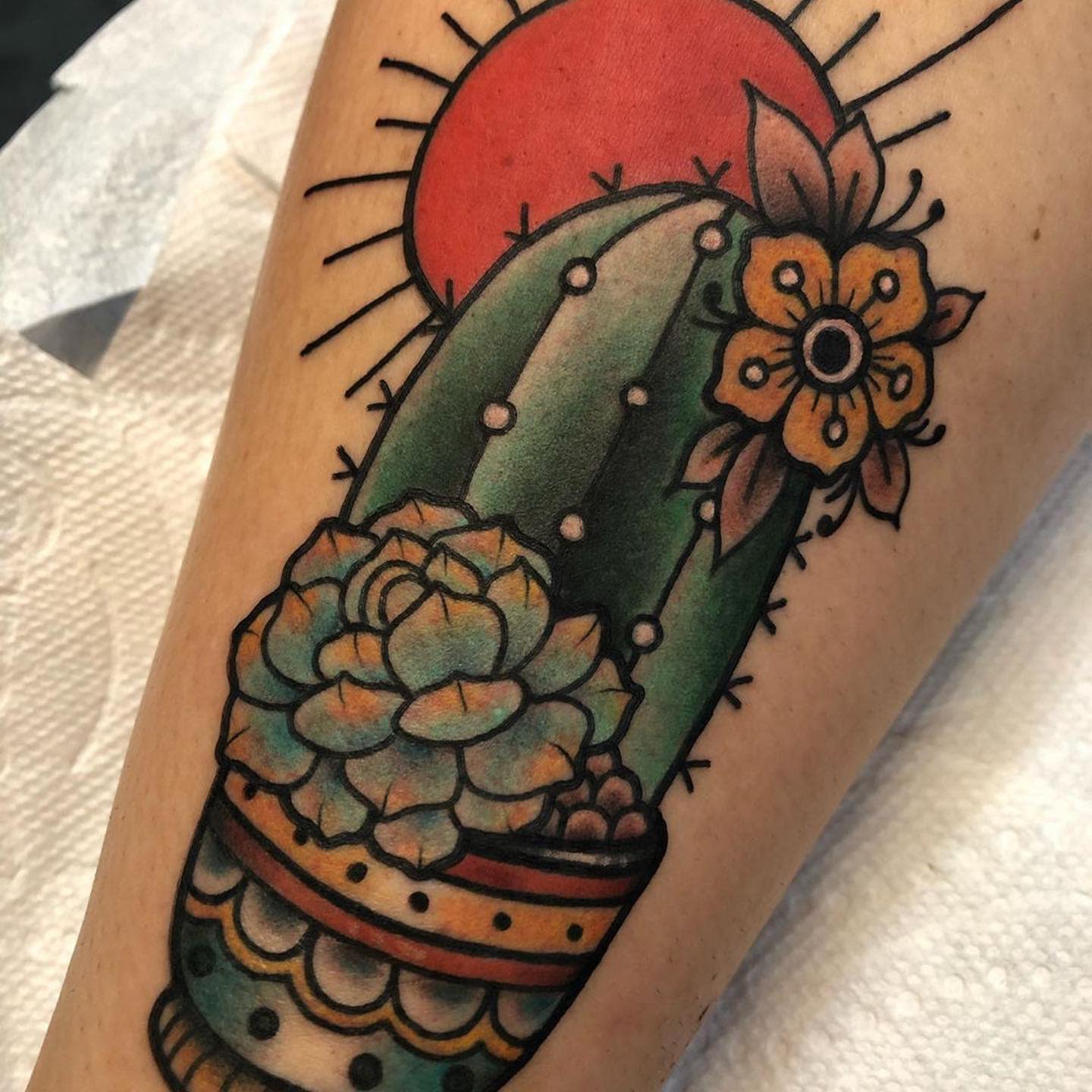 Tattoo featuring a cactus, succulents, and flowers in a pot with a red sun in the background