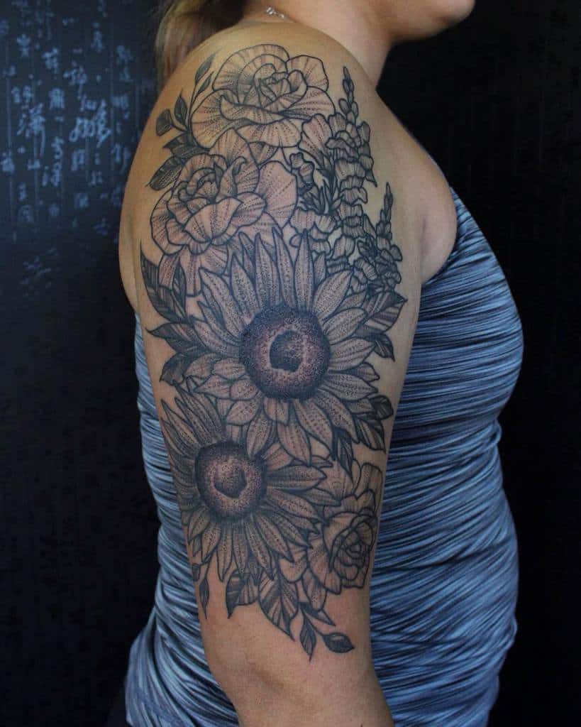 Black floral tattoo on woman's upper arm, featuring sunflowers and roses