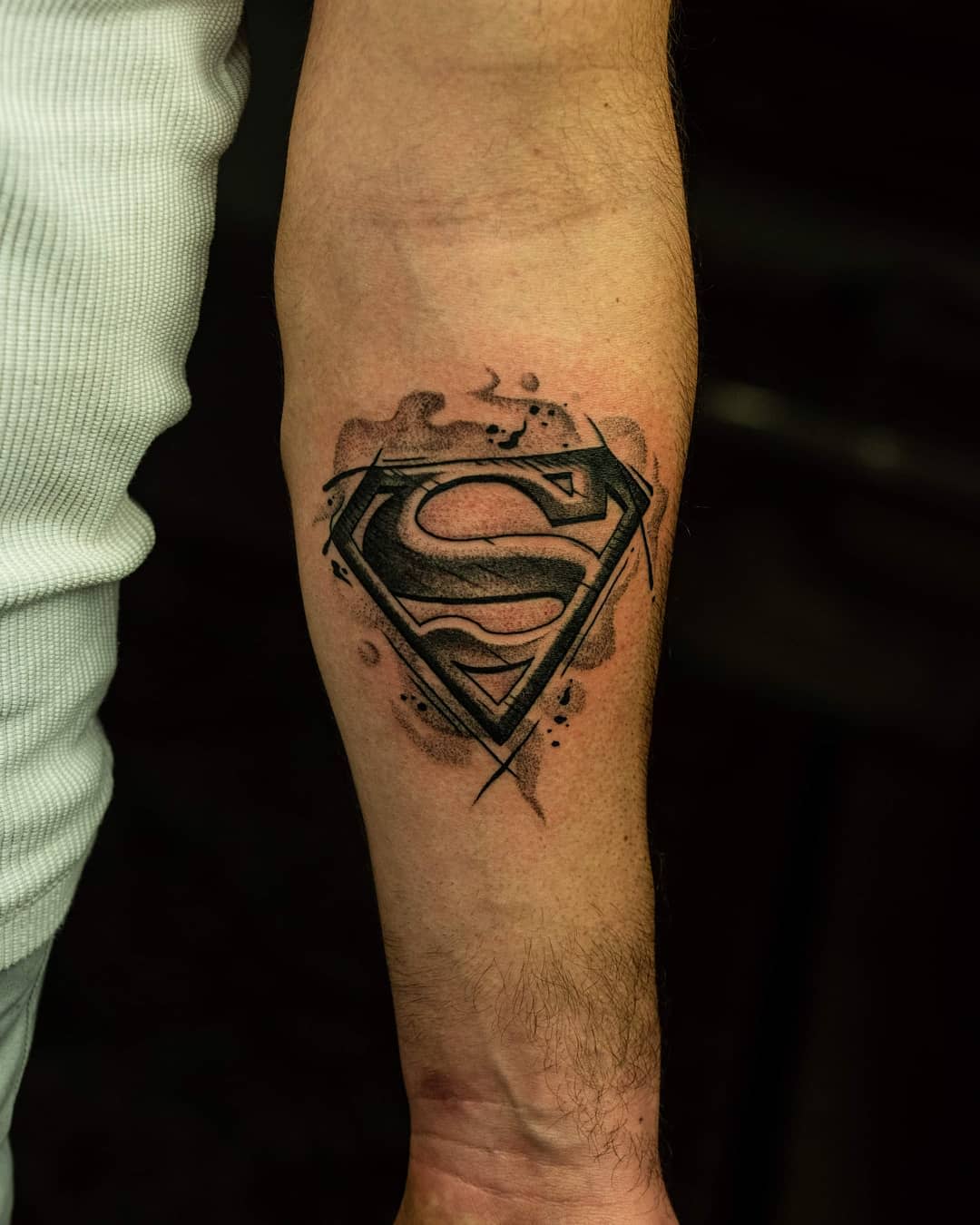 Forearm tattoo of Superman's "S" shield logo with artistic shading and splatter effect
