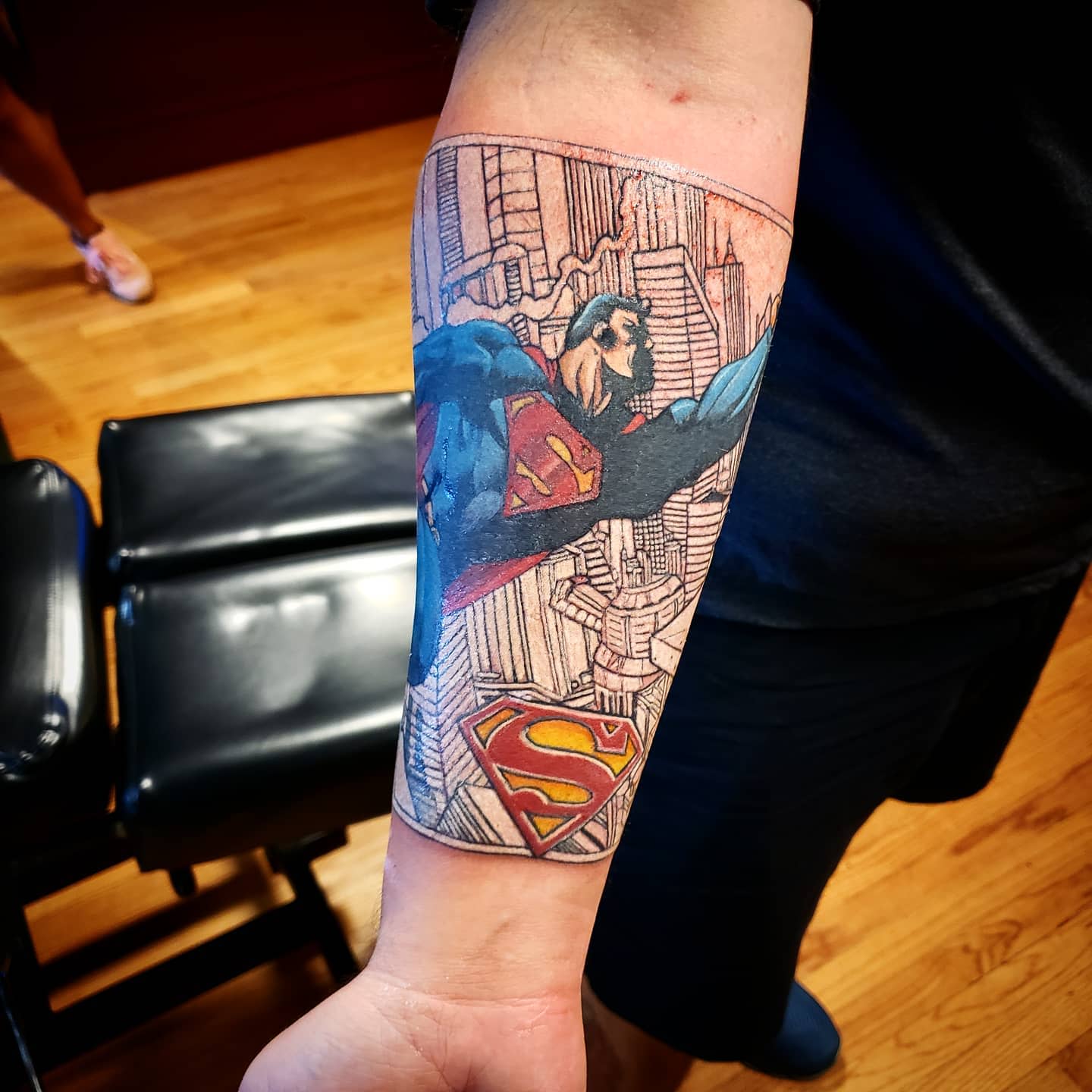 Forearm tattoo of Superman flying with a cityscape background and the Superman logo