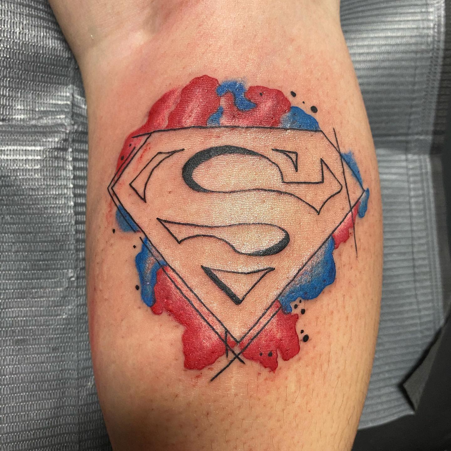 Superman logo tattoo with red and blue watercolor effects on a calf
