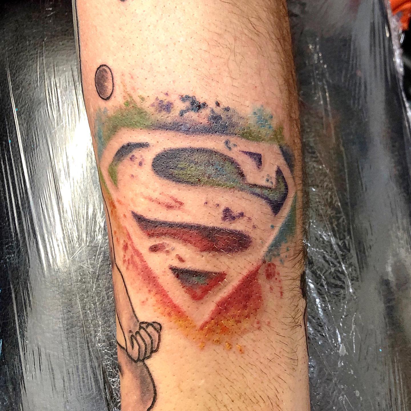 Forearm tattoo of the Superman logo with colorful splatter effects around it