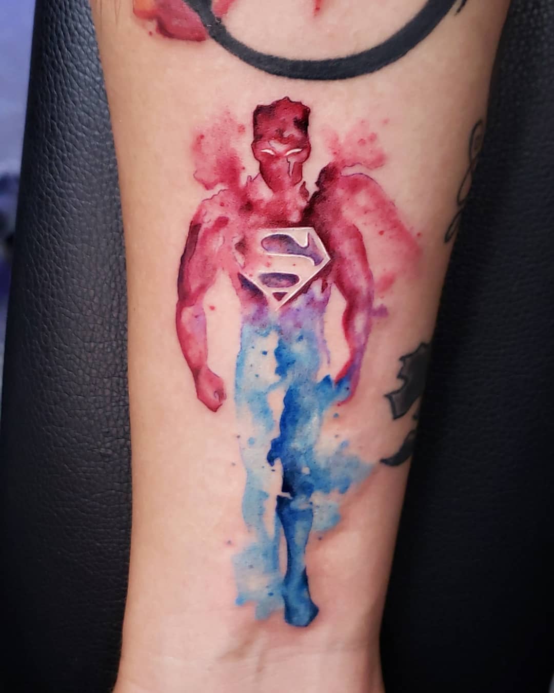 Watercolor tattoo of Superman in red, blue, and purple tones on a forearm