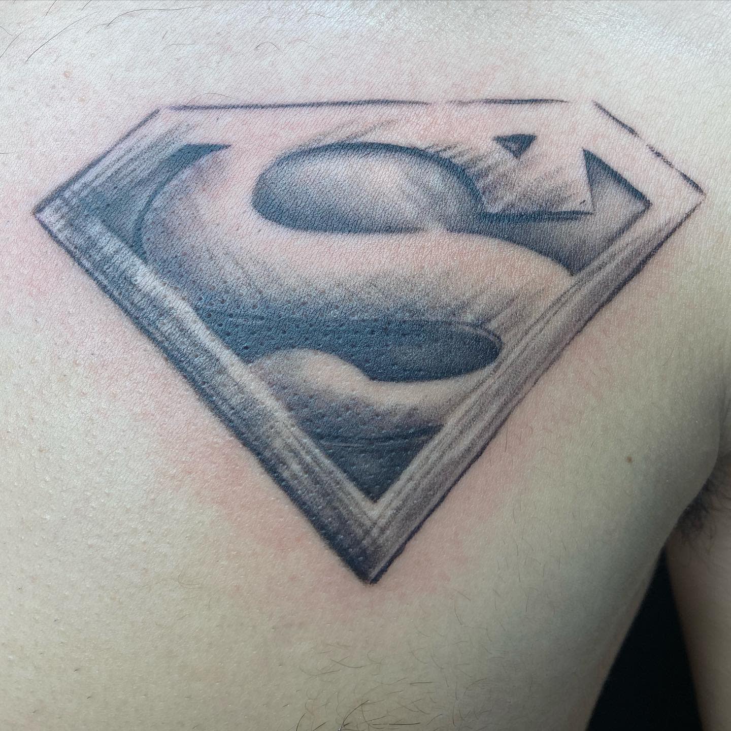 Black Superman logo tattoo on the upper chest, with shaded detailing and a 3D effect