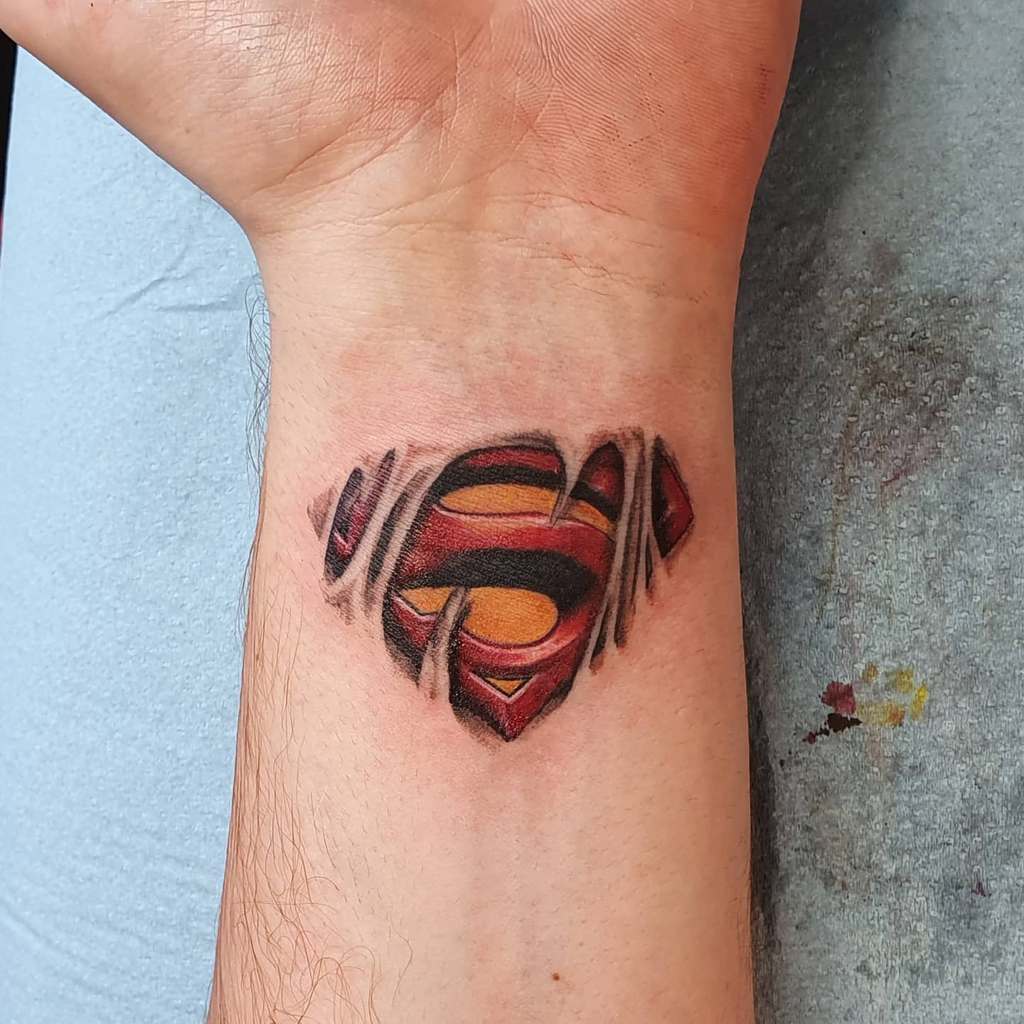 Tattoo of a torn skin revealing a Superman logo on a forearm